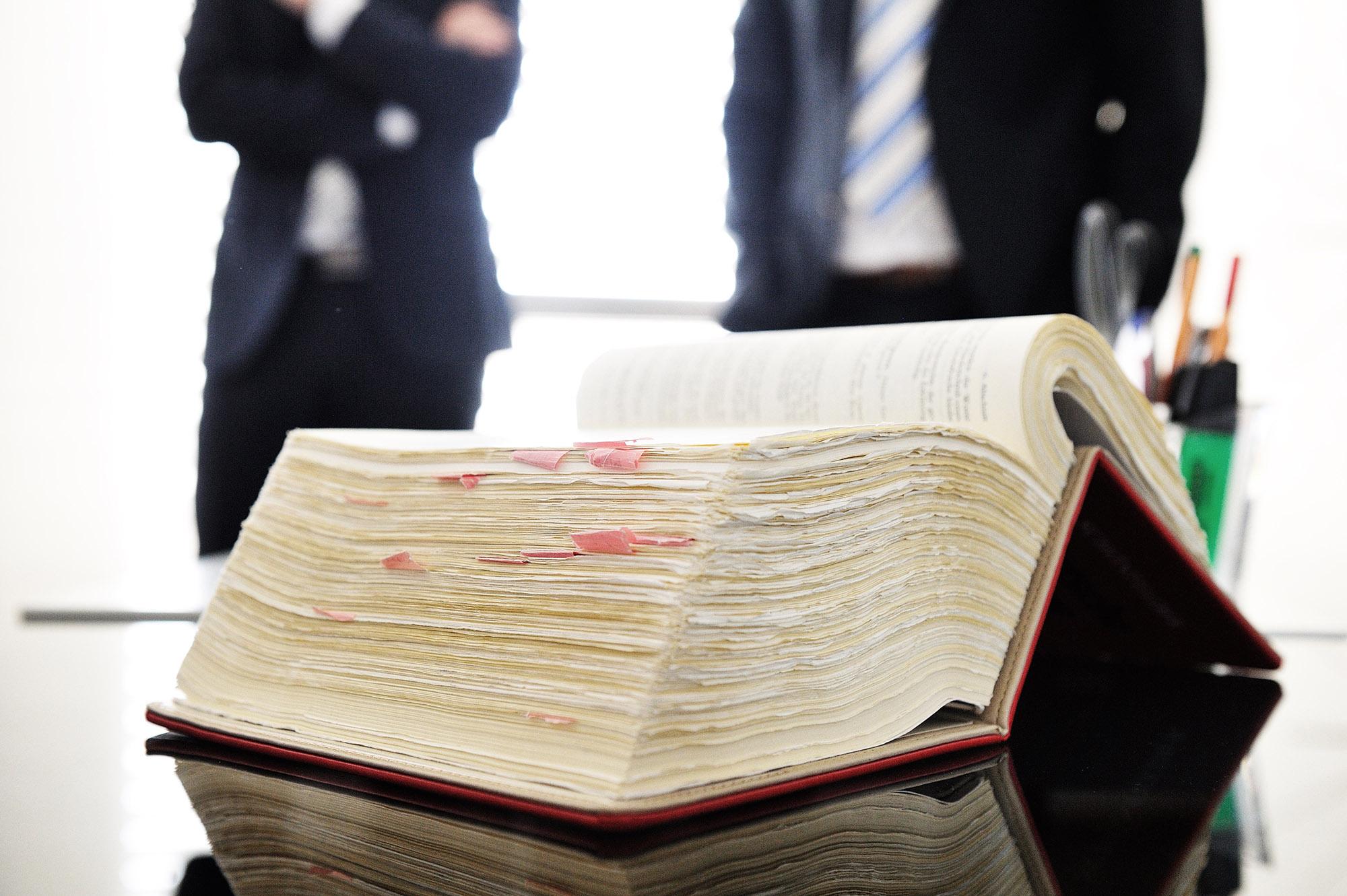 Dickes Buch mit Post-it's, dahinter stehen Menschen im Anzug