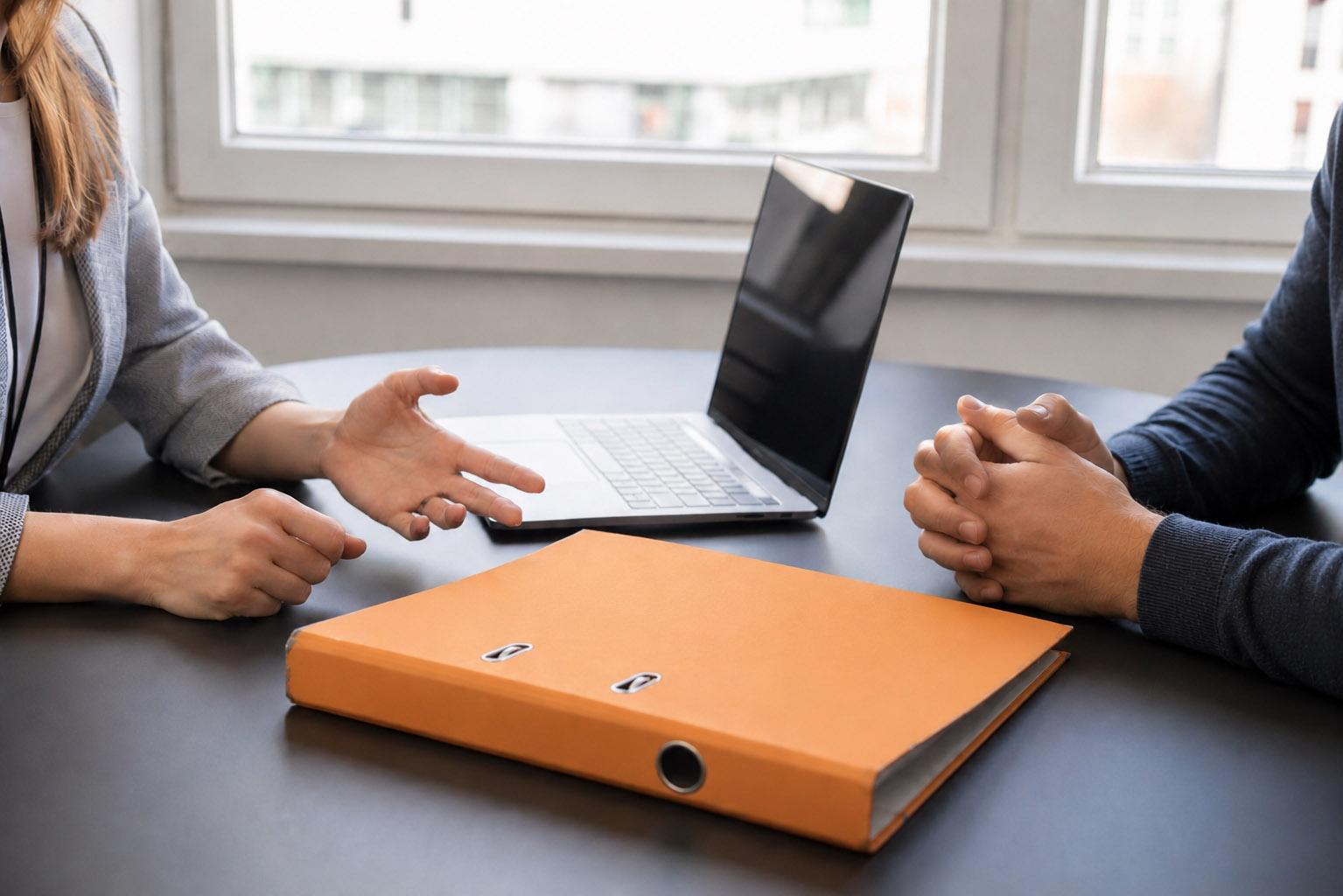 A woman in a business outfit and a man in a jumper are sitting opposite each other, with a folder lying beside them. Their faces are not visible; only their hands and upper bodies can be seen.&nbsp;