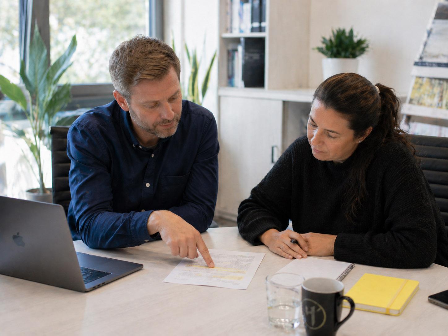 Two people are sitting at a desk, and one of them is pointing out a yellow mark on a sheet of paper to the other. A yellow notebook lies next to it.