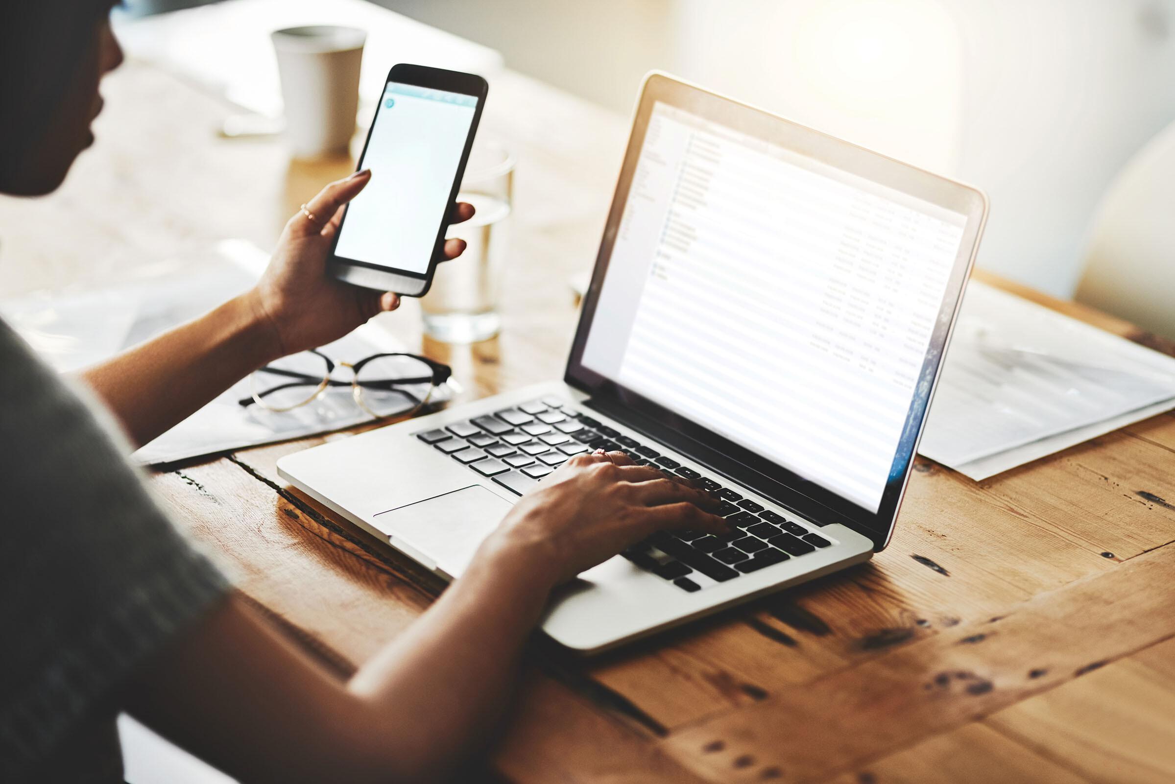 Eine Person arbeitet mit Handy und Laptop an einem Schreibtisch aus Holz