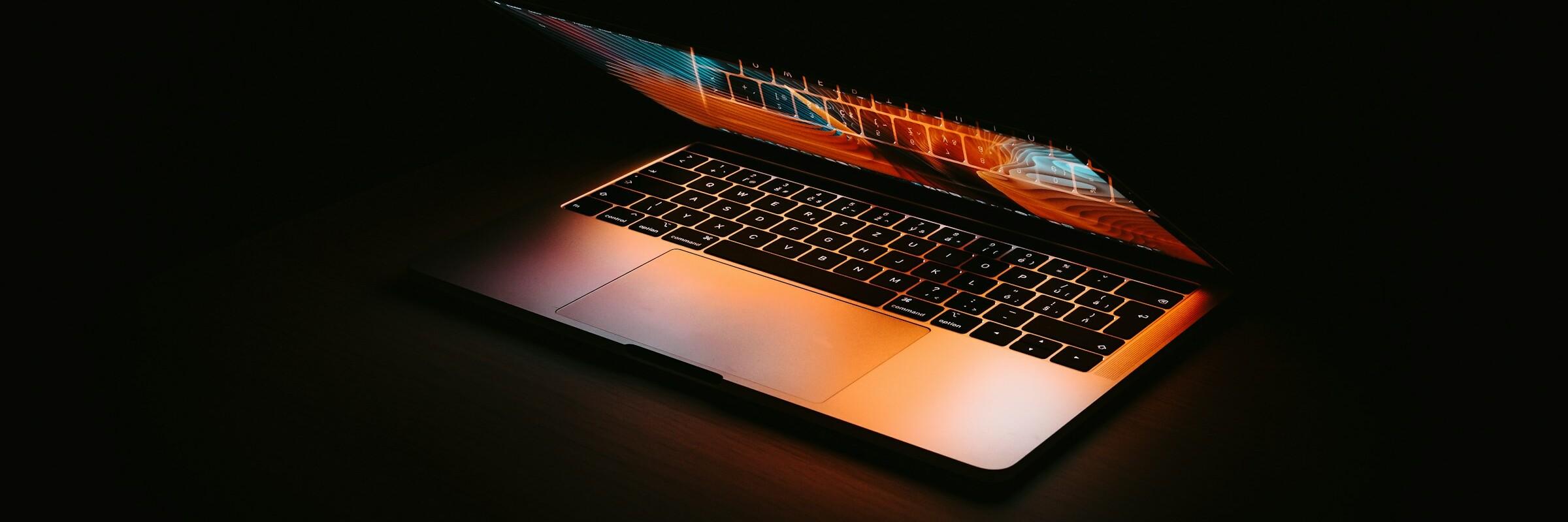 Aufgeklappter Laptop Die Farben vom Bildschirm eines halb aufgeklappten Laptops spiegeln sich auf seiner Tastatur; der Hintergrund ist dunkel. 