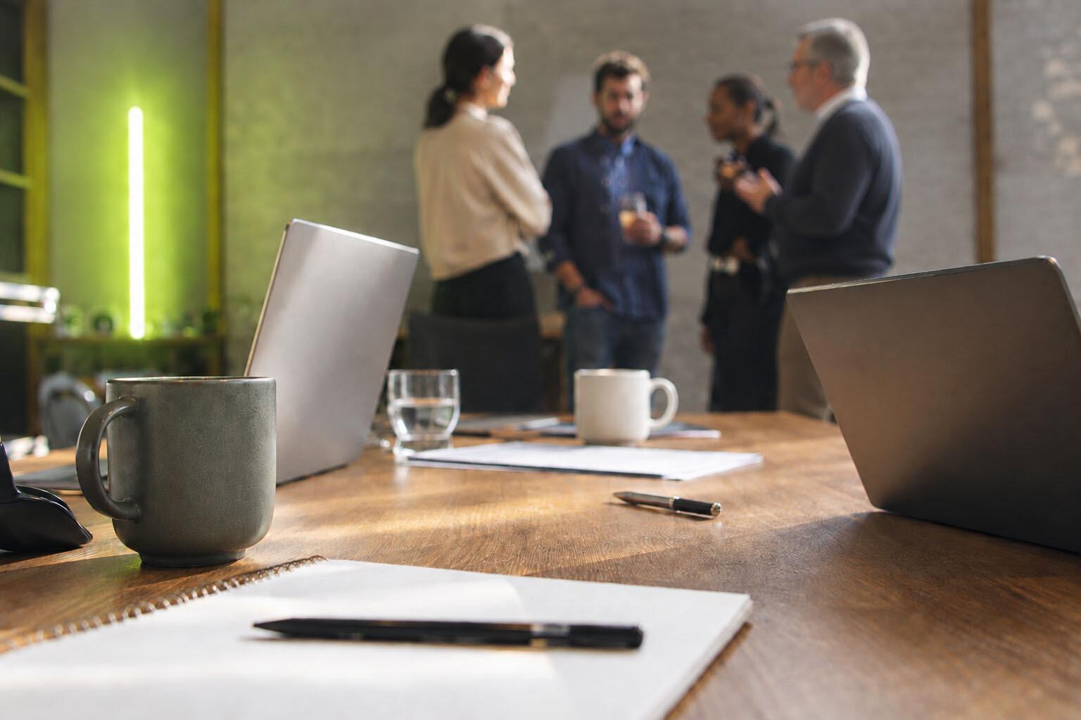 Networking Ein Konferenztisch mit Schreibmaterial, Laptops und Kaffeetassen, im Hintergrund stehen ein paar Personen im Gespräch zusammen