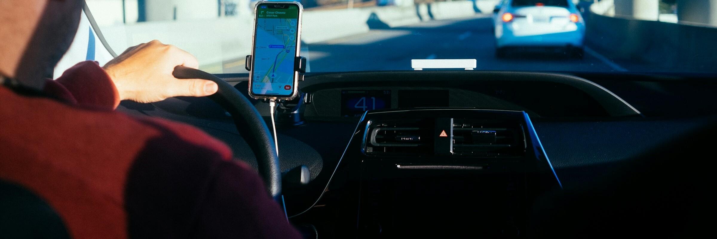 Man blickt einem Autofahrer vom Rücksitz aus über die Schulter; er blickt durch die Scheibe, vor ihm ist ein Auto und auf dem festgeklemmten Handy ist eine Navigations-App geöffnet.