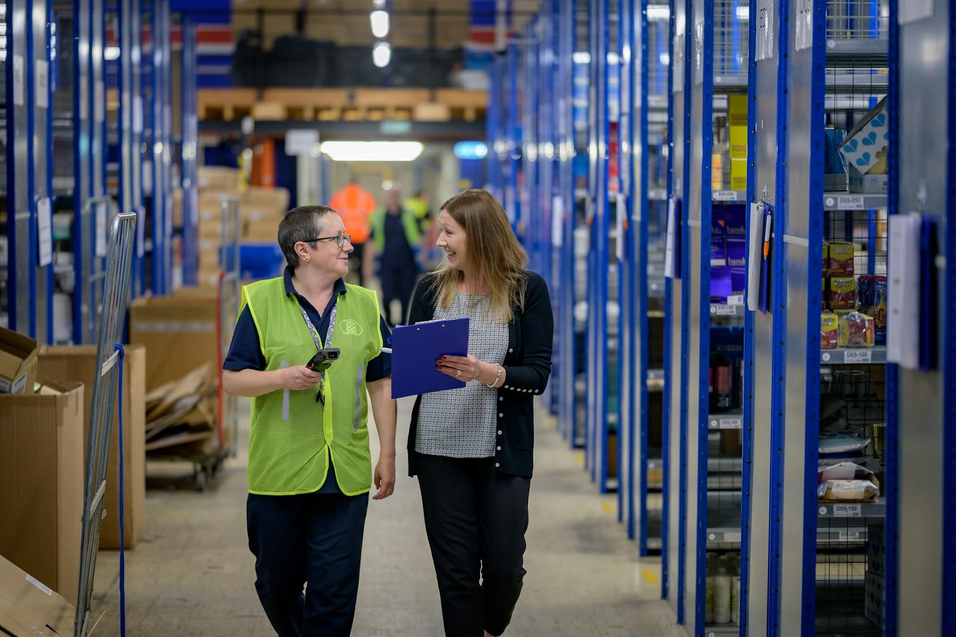 Zusammenarbeit Two women are walking through a warehouse; one is showing the other something on a clipboard