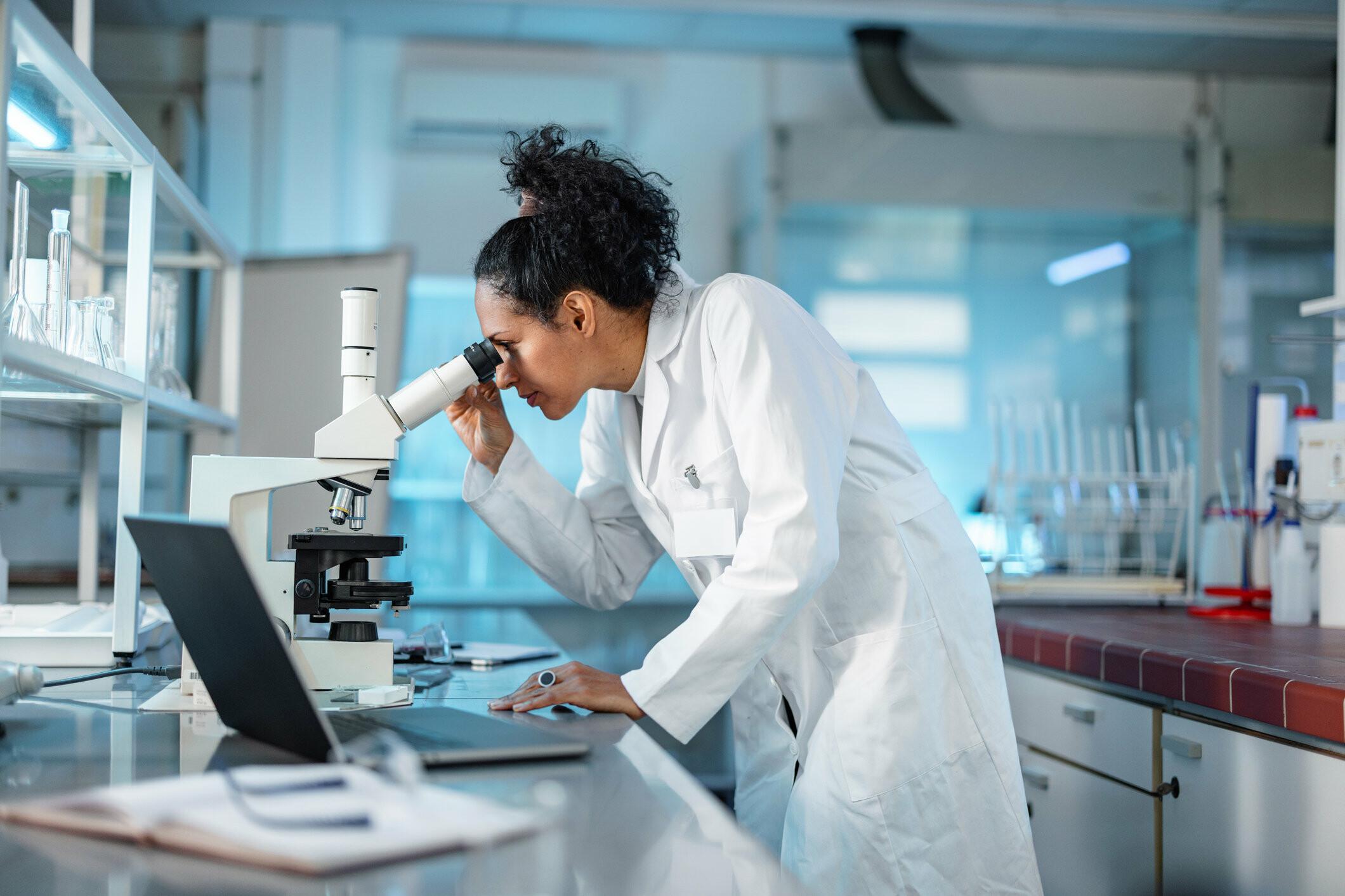 Frau im Labor A woman in a white coat looks at something through a microscope.