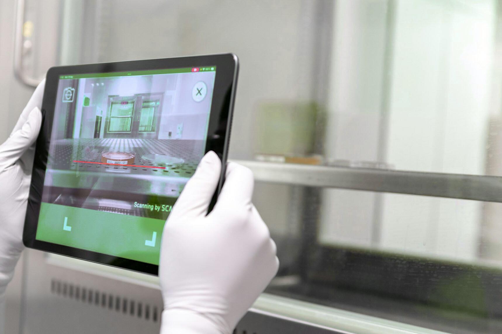 Check auf Tablet Zwei Hände mit Handschuhen halten ein Tablet und man sieht auf dem Bildschirm, wie ein runder Behälter gescannt wird.