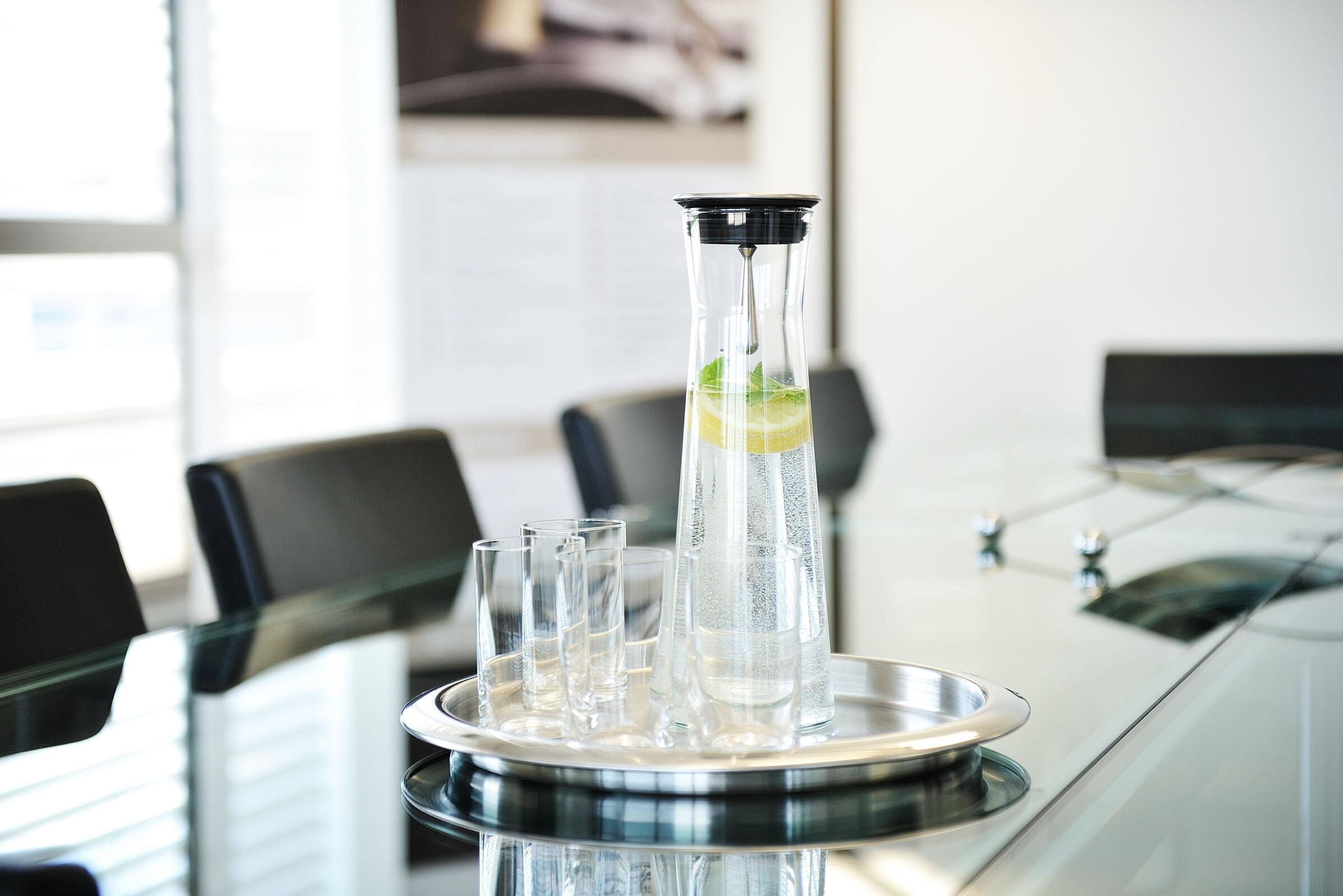 Glaskaraffe auf Glastisch A glass carafe containing water and a slice of lemon, accompanied by several glasses, on a tray placed on a glass table