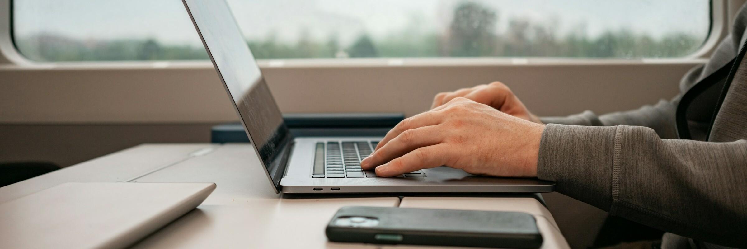 Mann mit Laptop mit Zug Die Hände eines Mannes arbeiten an einem Laptop, davor liegt ein Handy, im Hintergrund ist ein Zugfenster mit verschwommener Landschaft zu sehenEine männliche Hand hat einen Schlüssel in der Hand und dieser zeigt in Richtung eines Türschlosses